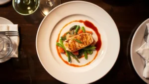 Overhead view of a perfectly plated fine dining dish with protein, seasonal vegetables, and artistic sauce drizzle on white ceramic plate, professional culinary presentation, warm restaurant lighting