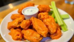 Crispy golden buffalo wings piled on a white plate with blue cheese dip in small ceramic ramekin, fresh celery sticks arranged beside, vibrant orange sauce glistening under warm restaurant lighting