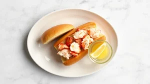 Overhead view of perfectly plated lobster roll on white ceramic dish with drawn butter in small glass bowl, fresh lemon wedge, and crispy toasted brioche bun, minimalist presentation on marble countertop
