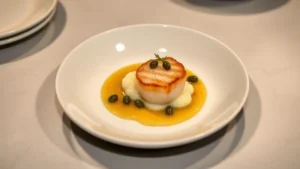 Overhead view of an elegantly plated fine dining dish with seared scallop, cauliflower purée, capers, and brown butter sauce on white porcelain, photographed in professional kitchen lighting with soft shadows
