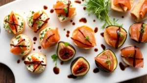 Overhead shot of elegant catering appetizer platter featuring herb-crusted goat cheese bites, crispy wontons, prosciutto-wrapped figs, and smoked salmon canapés arranged artfully with microgreens and balsamic reduction drizzle on white ceramic serving board